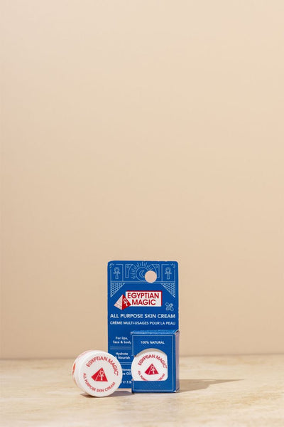 A small white jar labeled 'Egyptian Magic' sits inside a blue cardboard package with text that read Egyptian Magic, All Purpose Skin Cream with a translation in French underneath. Further text reads 100% natural, For lips face & body, Hydrating and Nourishing. With a small white jar in the forefront on a light beige surface against a matching beige background. 