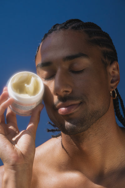 A close-up lifestyle shot of a man with braided hair holding an open white jar of Egyptian Magic near his face against a solid blue sky background.