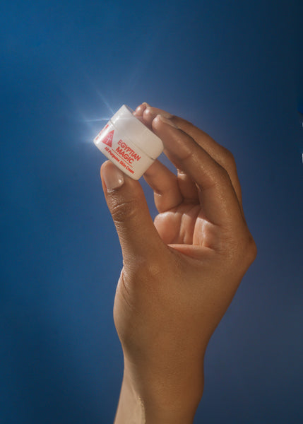 A hand holding a small .25oz jar of product with red text that reads 'Egyptian Magic All Purpose Skin Cream' against a dark blue background.